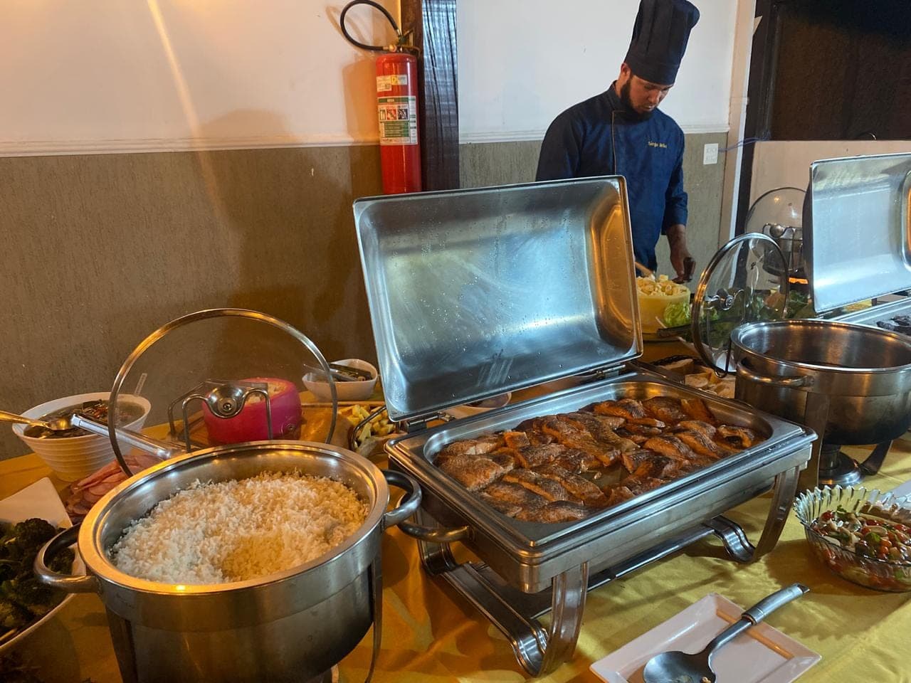 Rechauds abertos na linha do buffet, com o Chef ao fundo finalizando o serviço